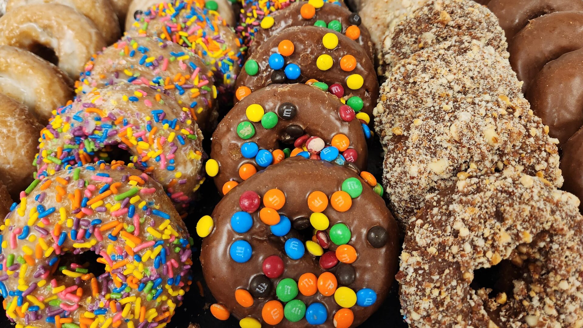 A colorful assortment of donuts arranged in rows, featuring a variety of toppings. Some are glazed, others are coated in rainbow sprinkles, mini M&M candies, crushed nuts, or chocolate icing. The vibrant and playful presentation makes them perfect for a festive breakfast or party treat.